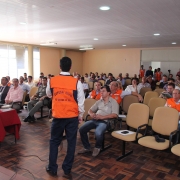 uma sala com várias pessoas sentadas observando uma palestra dada por um agente da defesa civil (ele usa uma jaqueta laranja da defesa civil) 