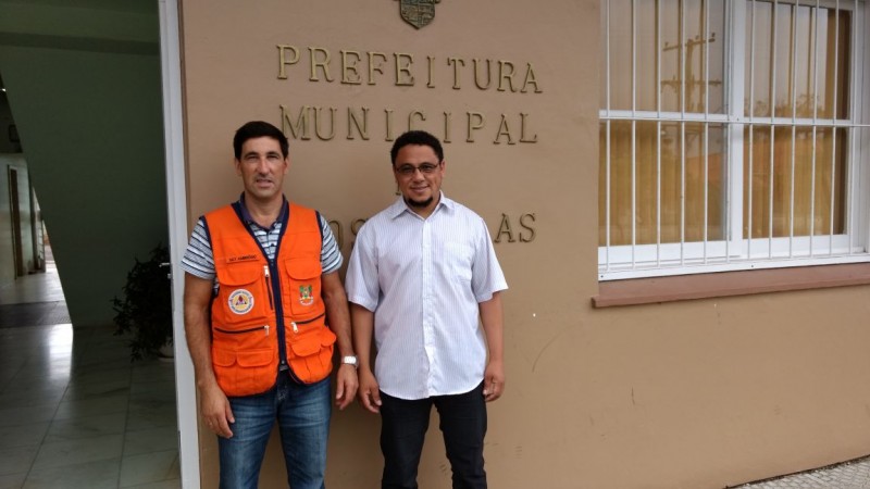 um coordenador da Defesa Civil está de colete laranja ao lado de um homem vestindo uma camisa branca. ambos estão posando na frente de uma parede onde está escrito "prefeitura municipal"