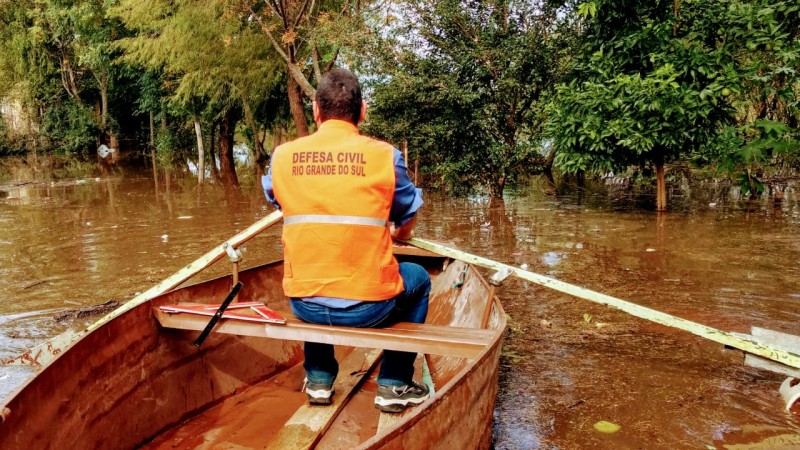 Um homem de colete laranja em uma canoa simples em meio a um rio com muitas folhas e com aspecto de sujo. Árvores completam o cenário, com a água quase em suas folhas