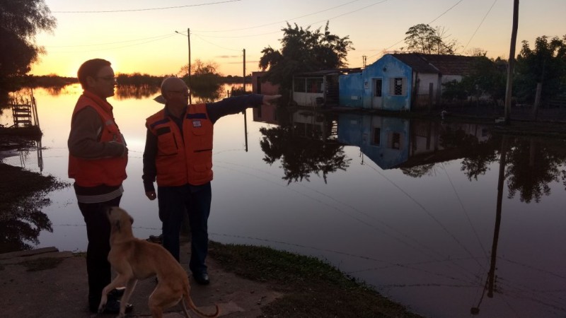 Dois homens analisando uma área residencial inundada. Um cachorro "estilo guaipeca" brinca com um dos homens, tentando chamar atenção. A área está realmente bem inundada.