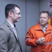 Dois homens conversando. Um usa terno claro e o outro uma jaqueta laranja