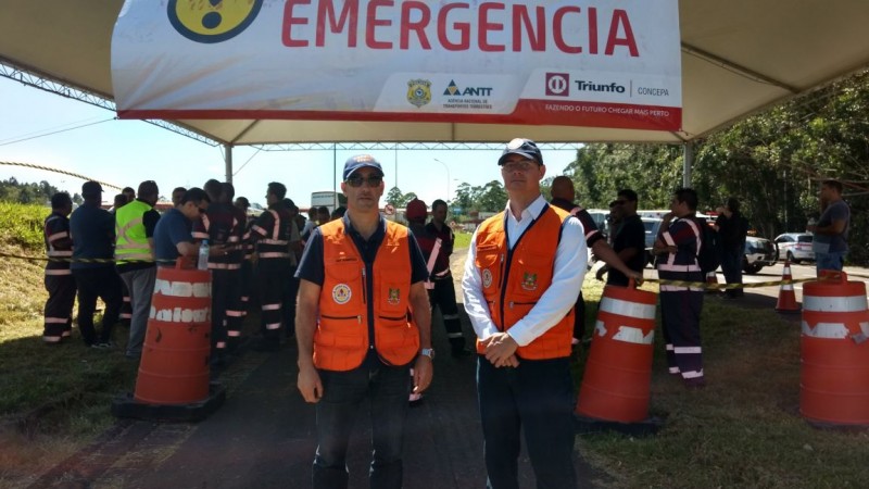 Dois homens de colete laranja, posando em frente a uma faixa escrito emergência, com outras pessoas fardadas atrás.