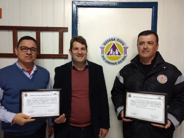Três homens em pé  com roupas escuras. Dois deles estão segurando certificados de reconhecimento e agradecimento.
