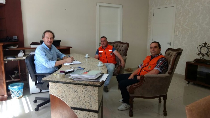 A foto foi tirada da lateral da mesa de reunião, onde se encontra de um lado o prefeito e do outro, os dois coordenadores da Defesa Civil. A sala tem as paredes claras, há duas portas ao fundo que estão fechadas.