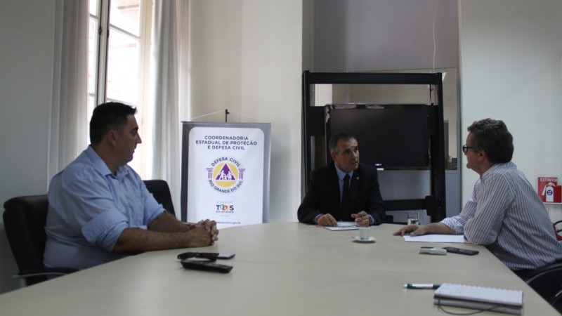 Uma sala de reuni&atilde;o com uma mesa grande de reuni&atilde;o. H&aacute; tr&ecirc;s homens em volta conversando