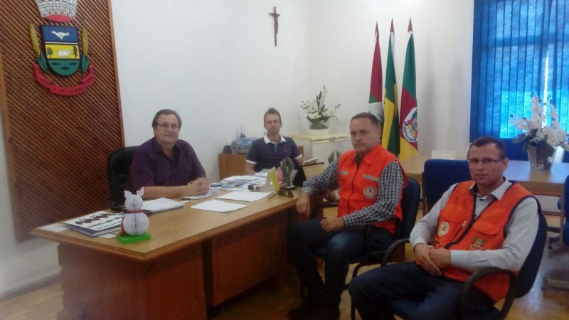 Quatro homens distribuídos por uma sala básica. Móveis de madeira clara, chão com textura clara. Paredes brancas e cortina azul.
