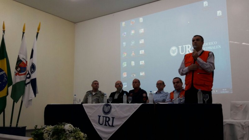5 homens sentados em frente a uma bancada enquanto um homem de colete laranja palestra para o público que não aparece na imagem