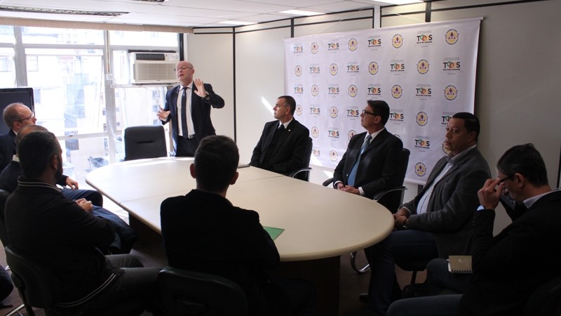 Mesa de reunião oval. Todos olham para a ponta. Um homem faz gestos enquanto fala. Todos usam roupa social. Ambiente bem iluminado e com clores claras.