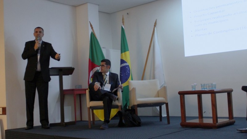 Um homem de preto (Coronel Avila) em pé em um palco falando ao microfone. Ao lado dele está sentado outro homem em uma poltrona. Atrás há algumas bandeiras, como a do estado e do Brasil. 