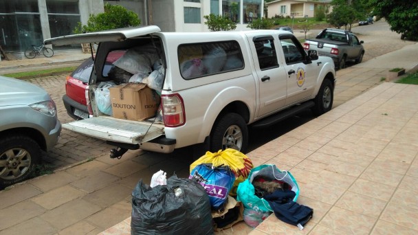 Uma caminhonete branca, com o logo da Defesa Civil, está com o porta malas aberto nele tem várias sacolas e caixas. Ao lado da caminhonete na calçada, tem mais sacolas com roupas.