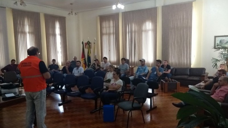 Um homem de laranja em frente a um público sentado. Fileiras mesclam cadeiras vazias e pessoas próximas. O ambiente parece pouco iluminado, porém as parede claras amenizam a situação.