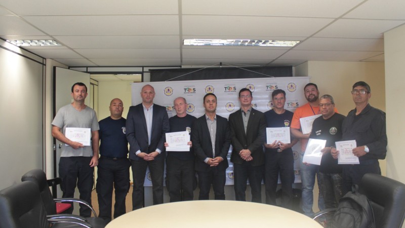 Dez homens em frente a um banner, segurando papeis em suas m&atilde;os. Um deles usa camiseta laranja, o resto veste roupas de tons escuros.
