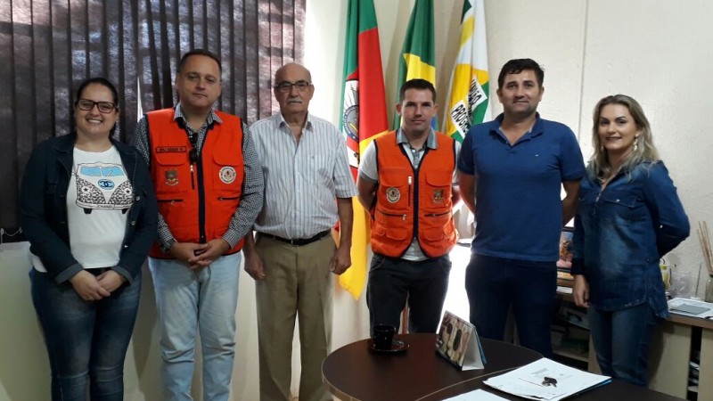 Seis pessoas em pé, com roupas claras. Ao fundo da sala, três bandeiras.