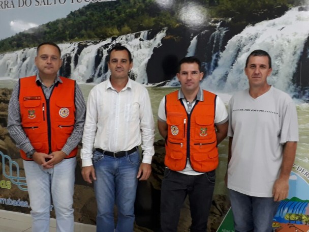 Quatro pessoas em pé, com roupas claras. Atrás dessas pessoas, tem uma foto de uma paisagem do município.