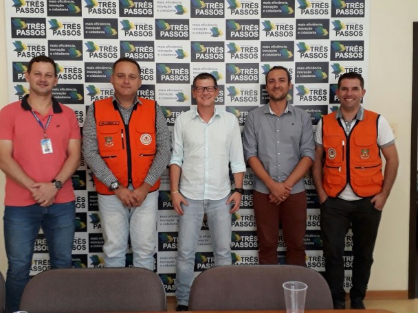 Cinco pessoas em pé com roupas claras. Ao fundo, tem um banner do município