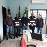 Cinco homens em pé, com roupas escuras. Uma sala bem iluminada e ao fundo bandeiras.
