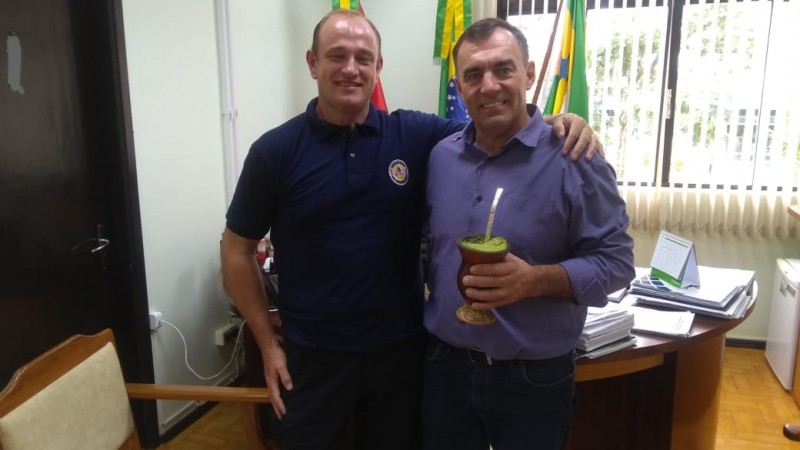 Dois homens em pé sorrindo pra foto, um deles segura uma cunha e chimarrão.