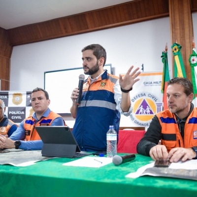 Gabriel Souza em pé atrás de uma mesa e ladeado por dois homens do lado esquerdo sentados e um do lado direito também sentado. Ele está com o microfone falando e gesticulando durante entrevista coletiva.