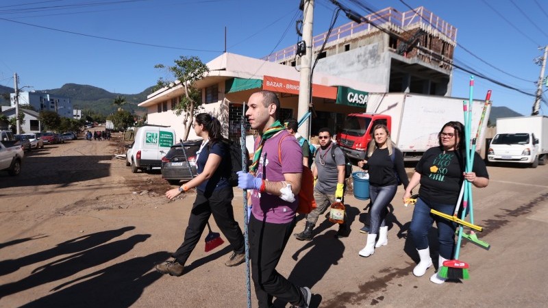 Voluntários caminhando pelas ruas carregando materiais para ajudar na limpeza de em uma das cidaddes atingidas pelas enchentes.