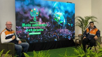 Os representantes da Defesa Civil estão sentados, com plantas em primeiro plano. Ao fundo, tela com slide sobre a Semana do Meio Ambiente