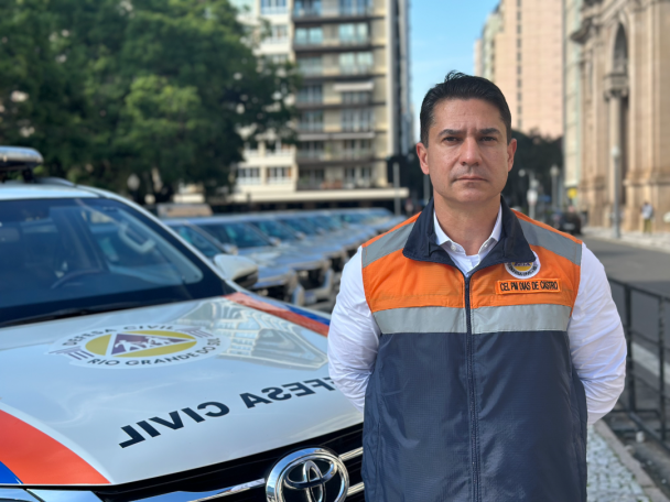Coronel PM Dias de Castro posa para foto em frente às novas viaturas. Ele está vestindo o colete da Defesa Civil do Estado.