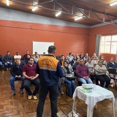 O palestrante, vestindo casaco da Defesa Civil, está de costas, falando para um público sentado