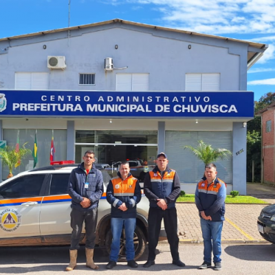 Quatro homens, três deles vestindo roupas da Defesa Civil, posam para foto na frente de uma viatura da Defesa Civil e da Prefeitura de Chuvisca