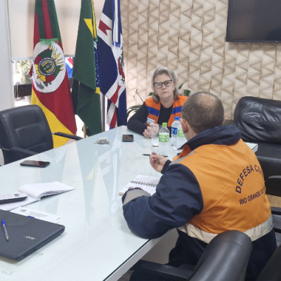 Três pessoas estão sentadas ao redor de uma mesa de reuniões conversando, uma está com jaqueta da Defesa Civil do RS e outra com colete de Defesa Civil municipal. As bandeiras do Brasil, do Rio Grande do Sul e de Dom Feliciano estão ao fundo.