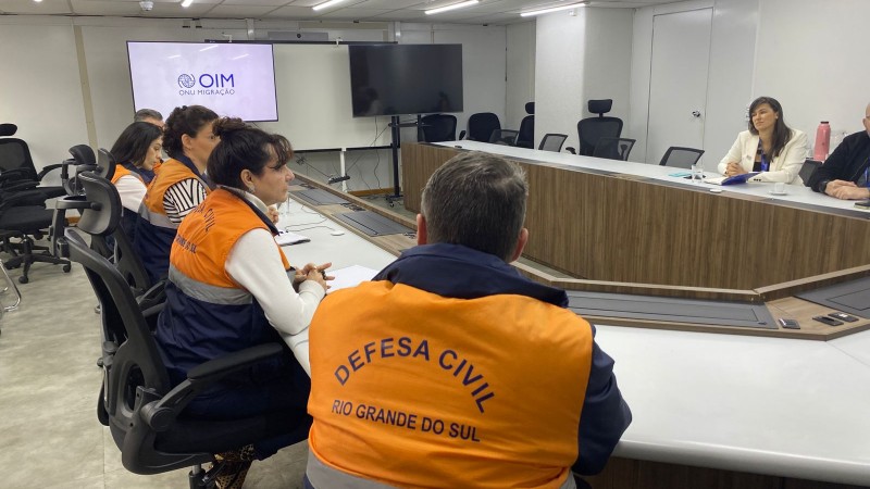 Pessoas est&atilde;o sentadas em mesa circular de reuni&atilde;o, algumas com colete da Defesa Civil. No fundo, uma tela aparece com a logo da OIM