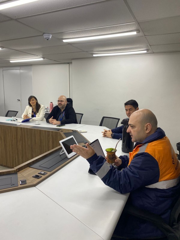 Pessoas estão sentadas em mesa circular de reunião, algumas com colete da Defesa Civil