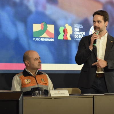 Governador Eduardo Leite se encontra de p&eacute;, com microfone na m&atilde;o. Sentados ao seu lado direito est&atilde;o coronel Boeira e coronel Dias.